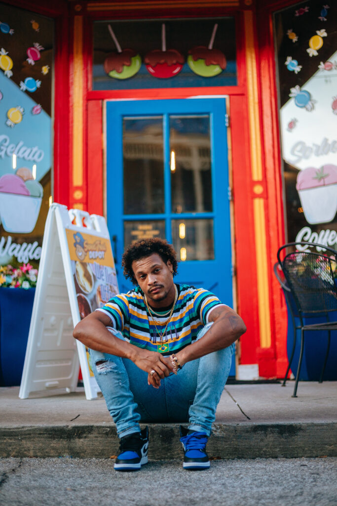 male model sitting on curb of a road beind a colorful storefront